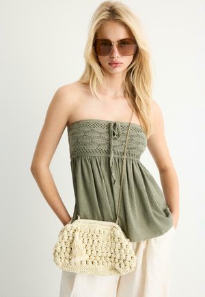 Plavuša koja nosi velike smeđe sunčane naočale, maslinasto zelenu top bez naramenica, bijele hlače, drži kremastu kvačkanu torbu s resicama.