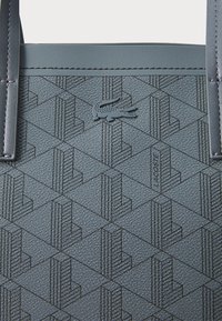 Črna teksturirana torba z geometrijskim vzorcem, ki vsebuje diskreten logotip Lacoste. Izdelana iz trpežnega materiala z dvema trdnima ročaji.