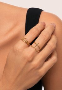 Gouden ringen met verschillende ontwerpen: één gladde band, één gebogen met een kruisaccent, getoond op een hand tegen een donkere top.