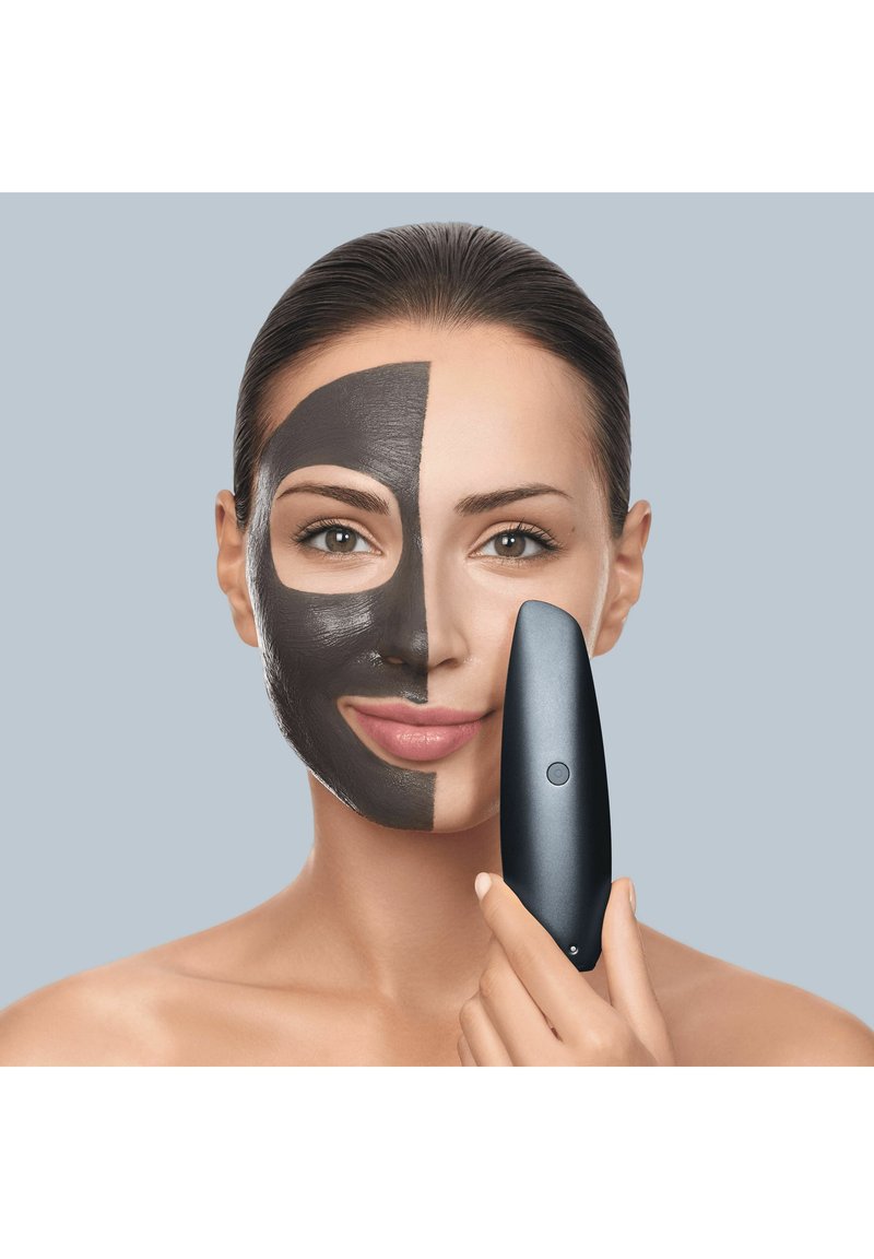 Kvinde med halvdelen af ansigtet dækket af en sort ansigtsmaske holder en elegant sort hudplejeenhed tæt på sin højre kind mod en ensfarvet baggrund.