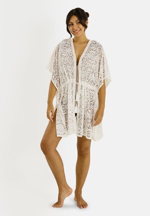 Femme debout pieds nus, portant une surcouche blanche en dentelle longueur genoux avec des manches larges et un lien devant, souriante, cheveux en chignon lâche.