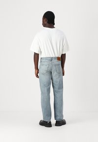 Light blue denim jeans with a relaxed fit, featuring a leather tag at the back waist and two back pockets. Worn with a white t-shirt and black shoes.