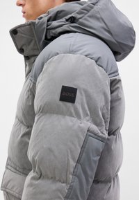 Chaqueta acolchada gris con capucha, que presenta una mezcla de texturas. Incluye un parche negro con el logo "BOSS" en la manga.