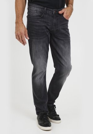 Homme portant un jean noir délavé et des baskets noires avec des semelles blanches, debout, une main dans la poche et l’autre main relâchée sur le côté.