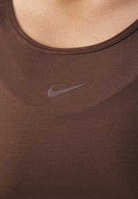 Brahna športna majica brez rokavov iz raztegljivega materiala, z okroglim izrezom in diskretnim logojem Nike v temnejši rjavi barvi na prsih.