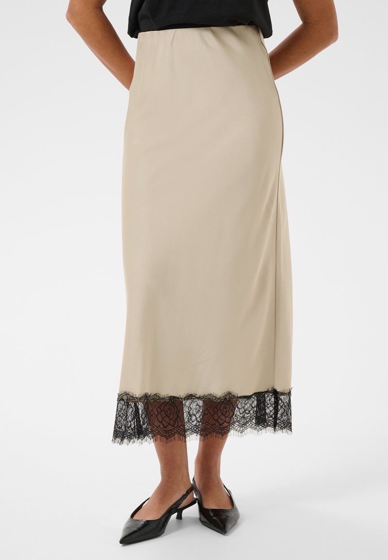 Jupe midi beige avec bordure en dentelle noire à l'ourlet, portée avec des escarpins slingback noirs à bout pointu et un top noir rentré.