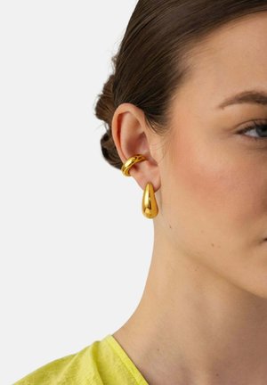 Junge Frau mit dunklem Haar trägt eine goldene Ohrmanschette am oberen Ohr und einen goldenen ohrenförmigen Tropfenanhänger am Ohrläppchen, heller Hintergrund.