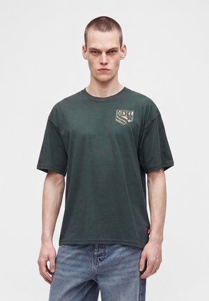 Mand iført en mørkegrøn Diesel t-shirt og lyseblå jeans stående mod en ensfarvet hvid baggrund.