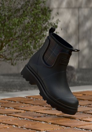Μαύρο αδιάβροχο μποτάκι αστραγάλου με καρό ελαστικές πλευρικές πιέτες, τοποθετημένο σε ξύλινα πλακάκια σε υπαίθριο χώρο κοντά σε φυλλώδες φυτό και τσιμεντένιο τοίχο.