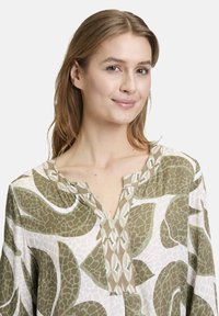 Lange mouw blouse van lichtgewicht stof. Heeft een geometrisch groen en wit patroon. V-hals met een subtiele kraagdetail.
