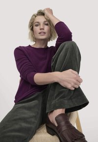Lila gestrickter Pullover, grüne Cordhosen und braune Slipper. Das Modell posiert sitzend mit einem angewinkelten Bein vor einem neutralen Hintergrund.
