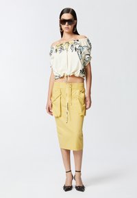 Bluzica s cvjetnim uzorkom u krem boji s suženim rubom. Žuta olovka suknja s dva prednja džepa i vezicom. Crne sandale s remenčićima.