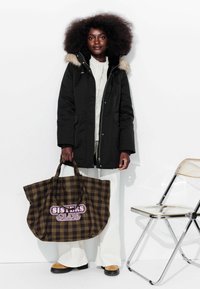 Parka noire avec capuche en fourrure, top crème, sac fourre-tout à carreaux avec "SISTERS CLUB" en rose, pantalon blanc évasé et chaussures beiges. Chaise transparente à proximité.