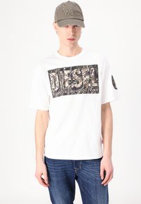 White t-shirt with a black and multicolored graphic panel that reads "DIESEL." Paired with blue denim jeans and a distressed gray cap.
