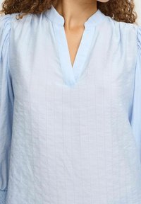 Blouse bleu clair avec un décolleté en V et des détails plissés sur les manches. Fabriquée à partir d'un tissu texturé, avec une coupe décontractée et des rayures subtiles.