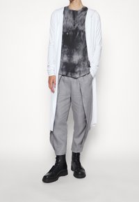 Lungo cardigan bianco sopra una maglietta a tinta unita nera e grigia, abbinato a pantaloni grigi chiari plissettati e stivali neri stringati. Design minimalista.