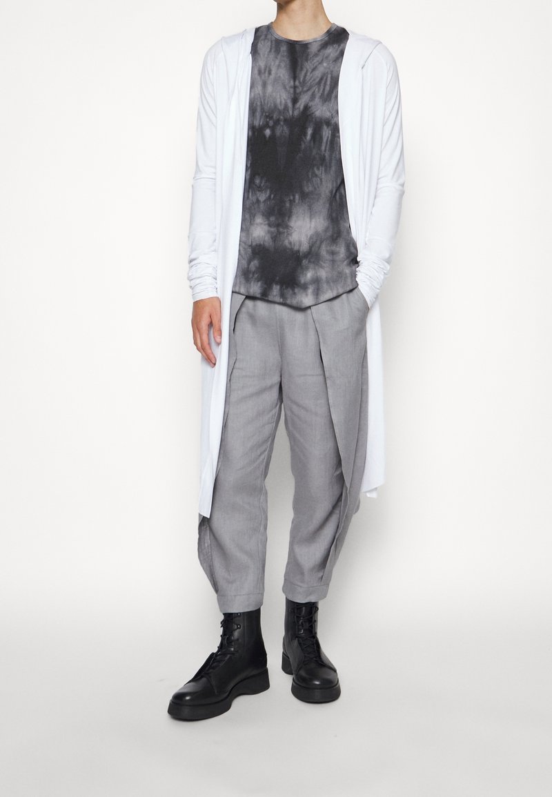 Lungo cardigan bianco sopra una maglietta a tinta unita nera e grigia, abbinato a pantaloni grigi chiari plissettati e stivali neri stringati. Design minimalista.