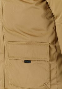 Giacca in tessuto khaki con texture diagonale, caratterizzata da una tasca a pattina rettangolare e un'etichetta nera sul bordo inferiore.