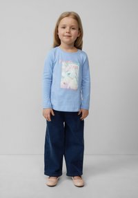Helles blaues Langarmhemd mit einem Einhorn-Graphic und Text, kombiniert mit dunkelblauen weiten Denim-Hosen und hellrosa Schuhen.
