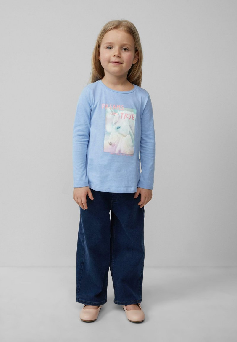 Helles blaues Langarmhemd mit einem Einhorn-Graphic und Text, kombiniert mit dunkelblauen weiten Denim-Hosen und hellrosa Schuhen.
