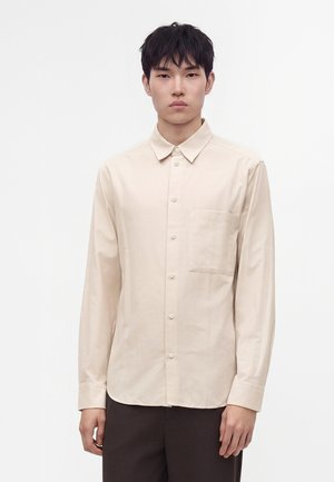 Jeune homme aux cheveux courts noirs, portant une chemise beige à boutons avec une poche poitrine et un pantalon foncé, sur un fond blanc uni.