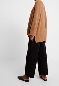 Beige ränderad tröja med långärmar, sidoflärpar, bärs över lösa svarta byxor och platta svarta skor. Enkelt mönster och tygtextur.