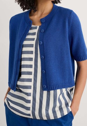 Femme portant un cardigan bleu à manches courtes boutonné sur un haut blanc avec des rayures verticales et horizontales bleu marine, mains dans les poches d’un pantalon bleu.