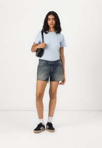 Lichtblauwe gebreide top met korte gerafelde mouwen, gecombineerd met versleten denim shorts en zwarte sneakers. Het model houdt een zwarte schoudertas vast.
