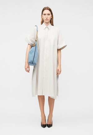 Femme portant une robe-chemise blanche ample arrivant aux genoux, avec un sac à bandoulière bleu clair à chaîne dorée, chaussée de talons noirs pointus, sur fond blanc.