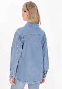 Vaaleansininen farkkutakki, jossa on kauluksellinen kaula, pitkät hihat ja napitetut hihansuut. Ominaisuuksiin kuuluu rento leikkaus ja tasainen, tasainen rakenne.