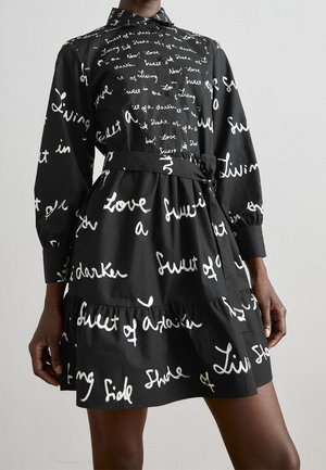 Schwarzes langärmeliges Kleid mit weißer, kursiver Schrift, tailliert mit Gürtel, getragen von einer Person mit dunklem Hautton vor einfachem Hintergrund.