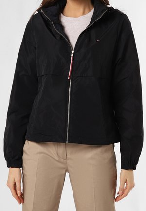 Femme portant une veste noire zippée avec des poignets élastiques et un pantalon beige, se tenant devant un fond uni.