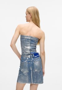 Ξανθιά γυναίκα φορώντας ένα strapless, μεταλλικής υφής τζην φόρεμα με πίσω τσέπες και ετικέτες Karl Lagerfeld Jeans, σε λευκό φόντο.