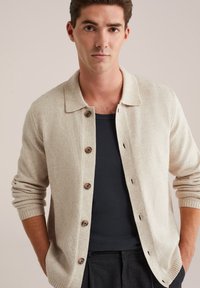 Cardigan bej tricotat, cu guler, cinci nasturi maro, mansete cu design în relief și o textură moale, purtat cu o cămașă închisă dedesubt.