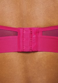 Sustentação rosa com painéis de rede, múltiplos fechos de gancho e olho, e uma textura suave, com letras da marca visíveis ao centro.