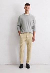 Mann steht an einer weißen Wand, trägt einen hellgrauen Sweater, beigetöne Hose mit teilweise in den Taschen versenkten Händen und schwarze Slipper.