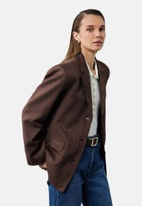 Brauner Wildleder-Blazer mit Reverskragen, Ein-Knopf-Verschluss und zwei vorderen Taschen, getragen über einem weißen Polohemd und blauen Jeans.