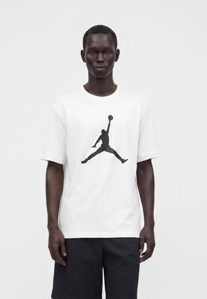 Homme portant un T-shirt blanc avec une silhouette noire d’un joueur de basketball en train de sauter, associé à un short noir, debout devant un fond uni.