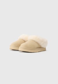 Une paire de pantoufles beiges en daim à enfiler avec une épaisse doublure en fourrure crème et des semelles en caoutchouc sur un fond blanc uni.