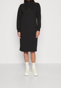 Schwarzes Langarmkleid mit hohem Kragen und Reißverschluss am Halsausschnitt, mit geradem Schnitt und seitlichen Schlitzen für mehr Bewegungsfreiheit.
