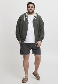 Siva zip-up dukserica s svijetlim unutarnjim oblogom, bijela majica, tamnosive kratke hlače i smeđe sandale. Dizajnirano za udobnost, ležeran kroj.
