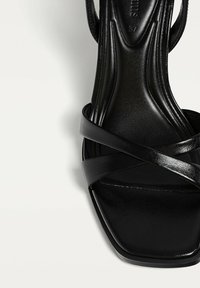 Schwarze High-Heel-Sandale aus glattem Leder mit überkreuzten Riemen und quadratischer Zehenpartie. Texturierte Innensohle und schlanke Silhouette.