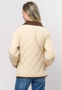 Beežist tikitud jakk pruuni sametkraega, millel on rombimuster ja elastsed varrukasäärised. Kandmisel siniste teksadega, vaadatuna tagant.