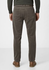 Mann in grauen Cordhosen, braunen Lederschuhen und einem dunklen Langarmoberteil, von hinten vor einem schlichten Hintergrund gezeigt.