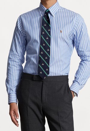 Homme portant une chemise à boutons rayée bleue et blanche avec un logo brodé, une cravate bleu marine à motif et un pantalon gris foncé, main dans la poche.