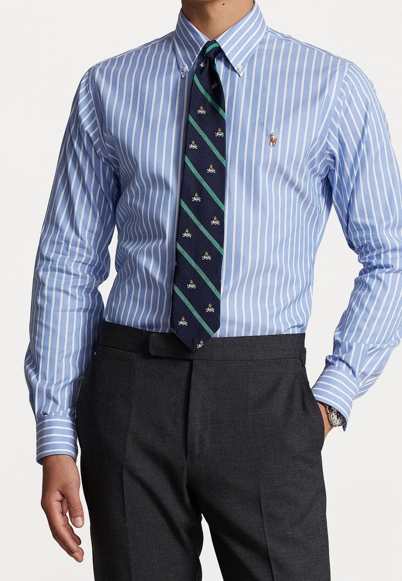 Homme portant une chemise à boutons rayée bleue et blanche avec un logo brodé, une cravate bleu marine à motif et un pantalon gris foncé, main dans la poche.