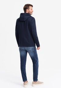 Marineblå hættejakke, blødt materiale, skræddersyet design, rygventilation, parret med lyseblå jeans og beige sneakers.