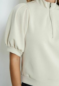 Light beige pullover with a high collar and half-zip closure. Features puffed sleeves and a fitted waistband, made from a soft, textured fabric.