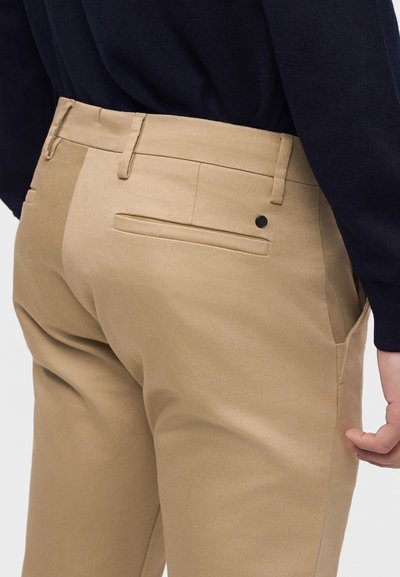 Beige bukser med bæltestropper og påsat lomme, båret med en mørk langærmet top på en person vist fra taljen og ned.