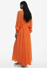 Lång orange chiffonklänning med avsmalnad midja, trumpetärmar och flödande kjol, med en lätt och genomskinlig textur.